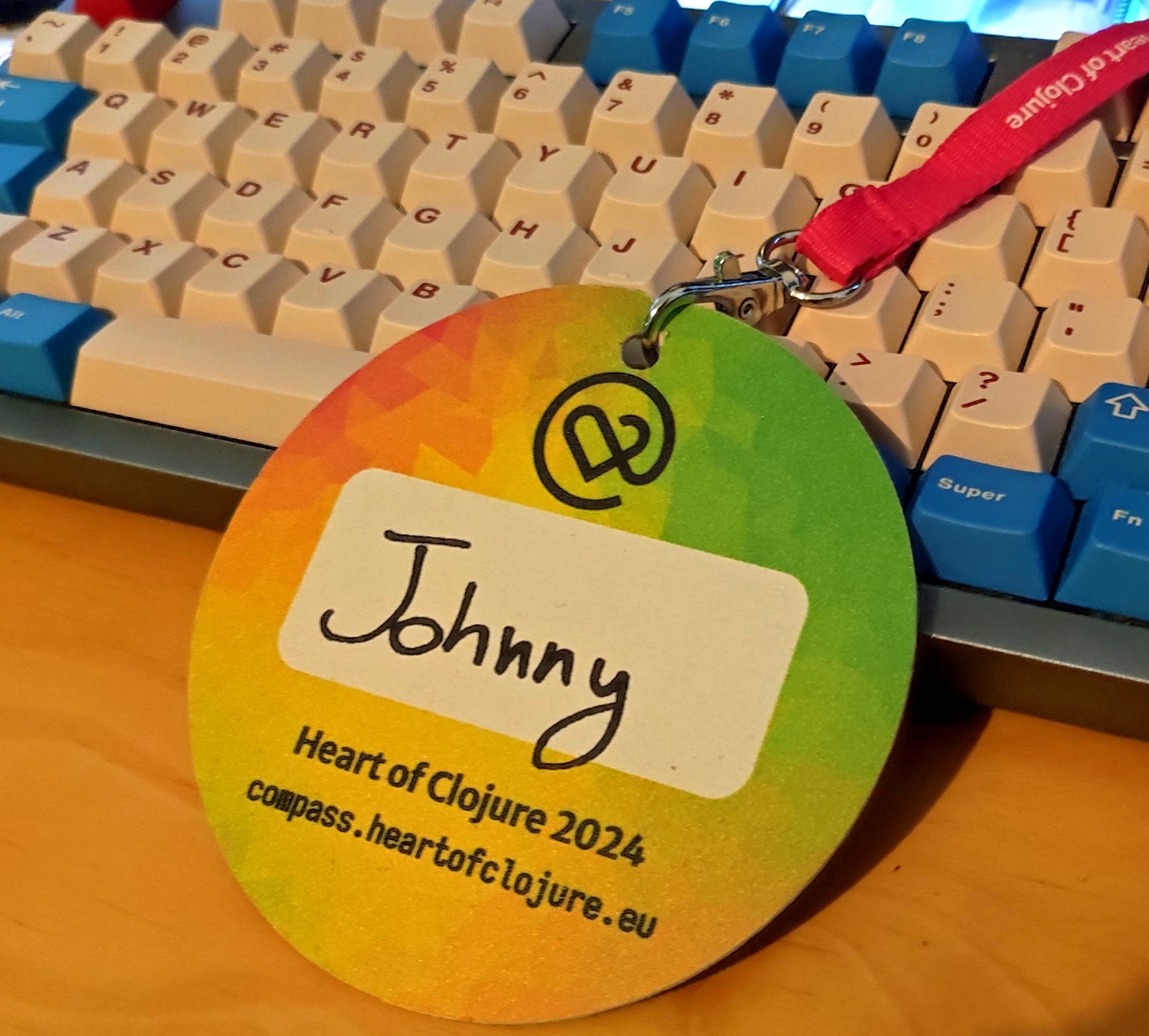 My Heart of Clojure badge, attached to a pink lanyard, leaning against my keyboard. The badge is a circle, with a red-yellow-green gradient pattern, the logo, a white text field with a handwritten "Johnny" on it, and the text "Heart of Clojure 2024 compass.heartofclojure.eu" below that.