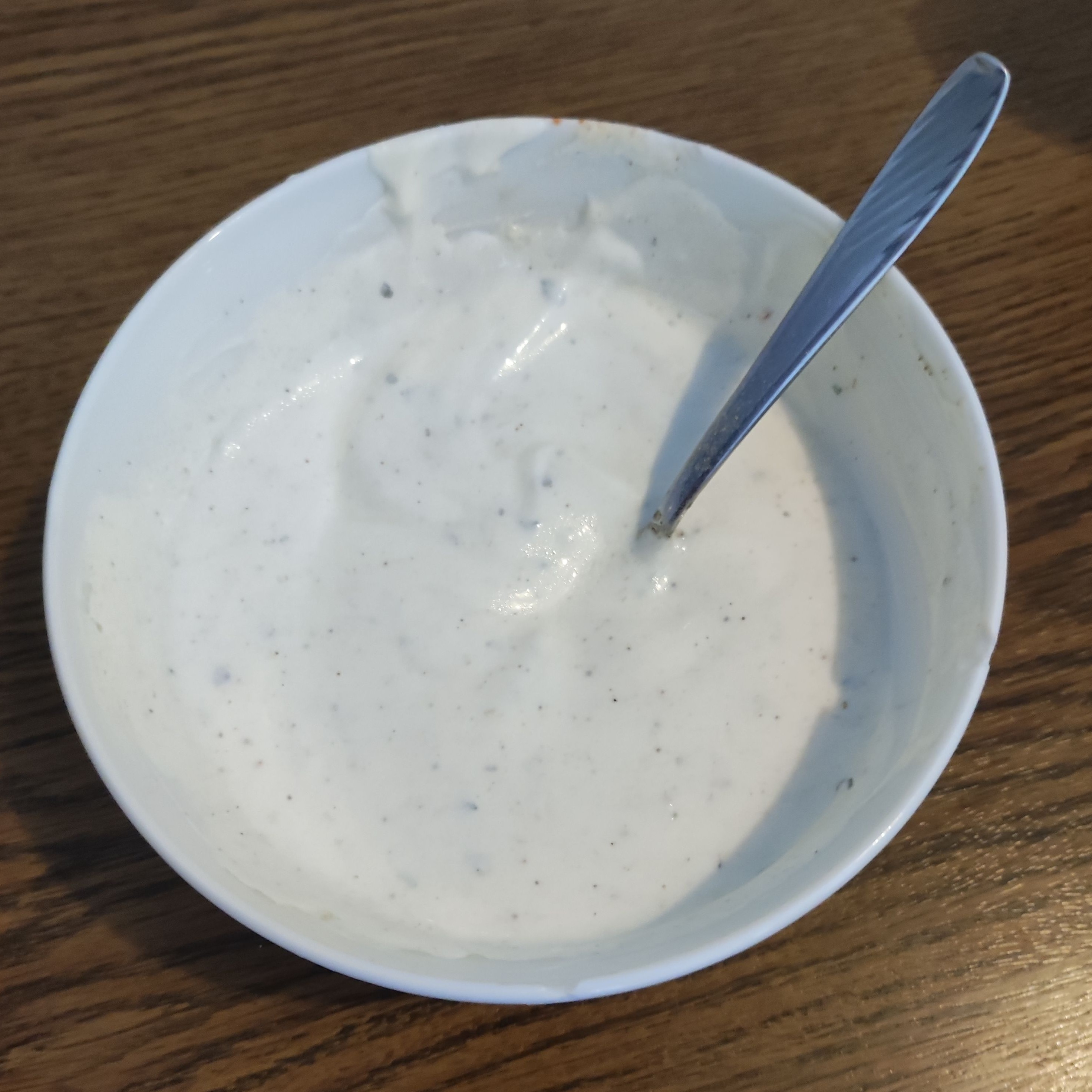 A small white porcellain bowl on a wooden table containing a yogurt-olive oil-lime mixture with spices. You can see particles of the ingredients in the otherwise white yogurt.