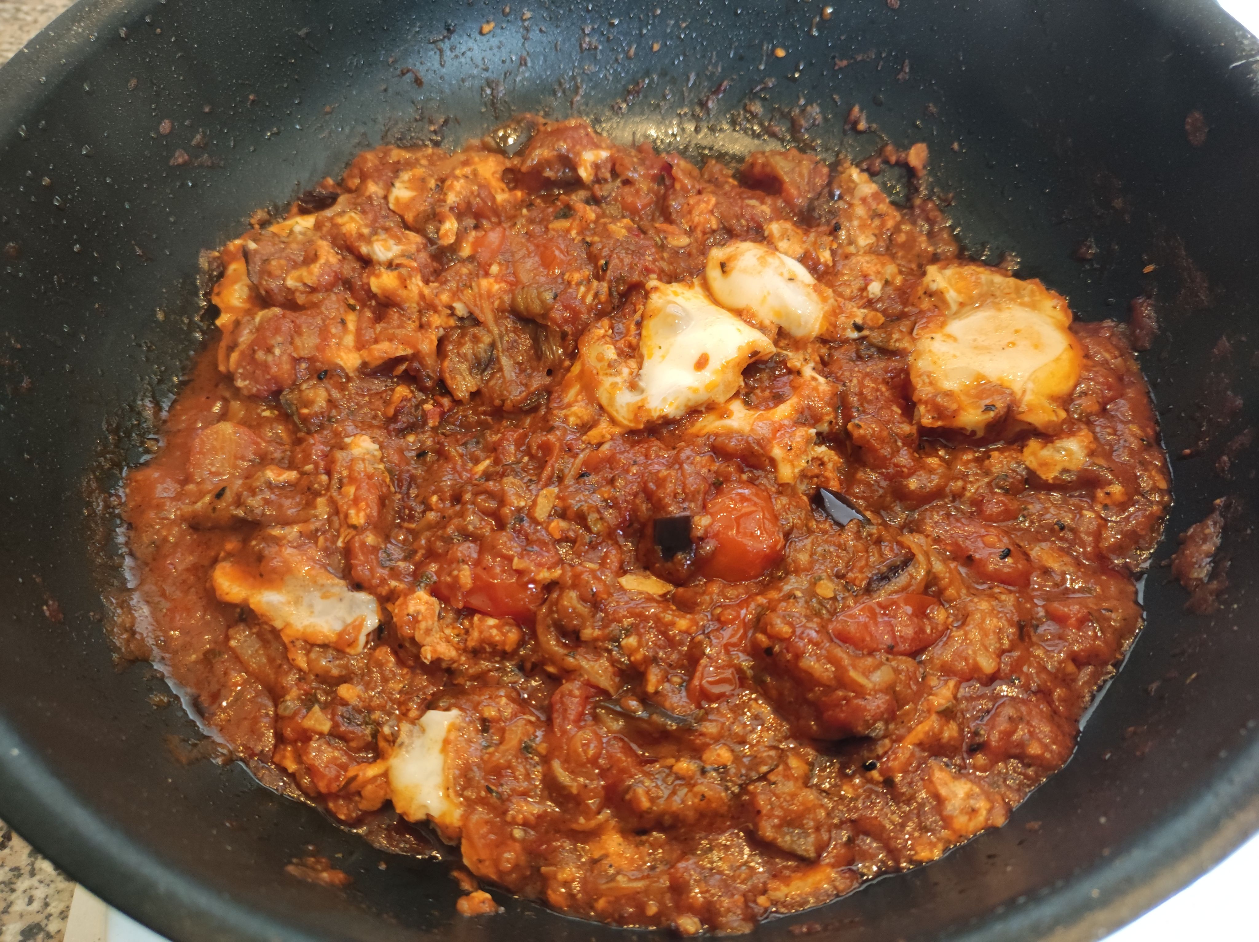 A deep non-stick frying pan containing some shakshouka. The cooked eggs have been broken up into smaller pieces and partially covered with the tomato sauce. You can see some fresh cherry tomatoes and little bits of black from eggplant pieces. Some raisins are also visible.
