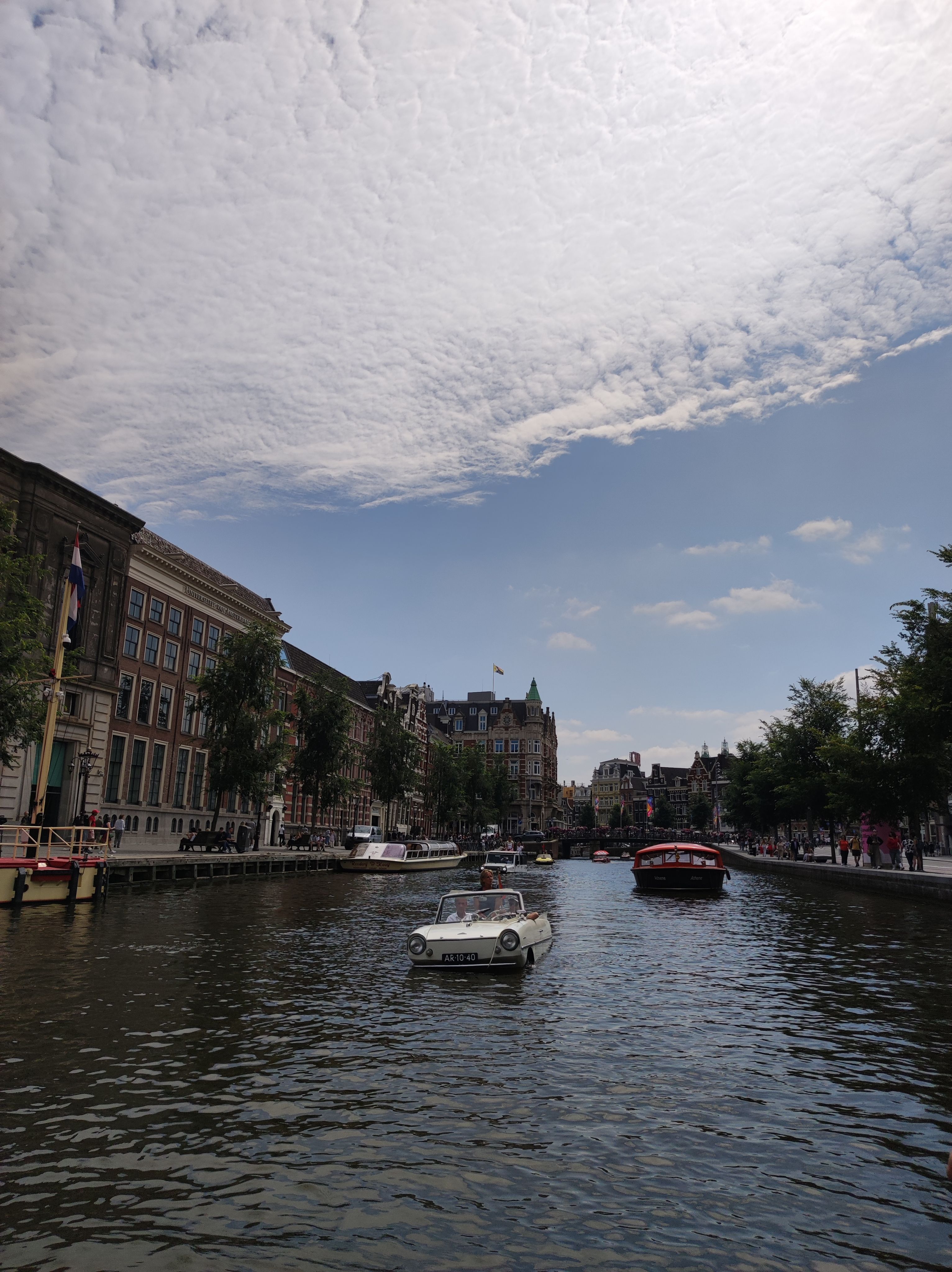 Photo taken on the canals of Amsterdam. We are on a rather wide canal, with pedestrian streets and trees left and right, and some 4 to 5 story buildings visible on the left side and in the background, behind a bridge. Directly facing us is a boat made from the skeleton of an old, white car. It looks like a car is driving in the water.