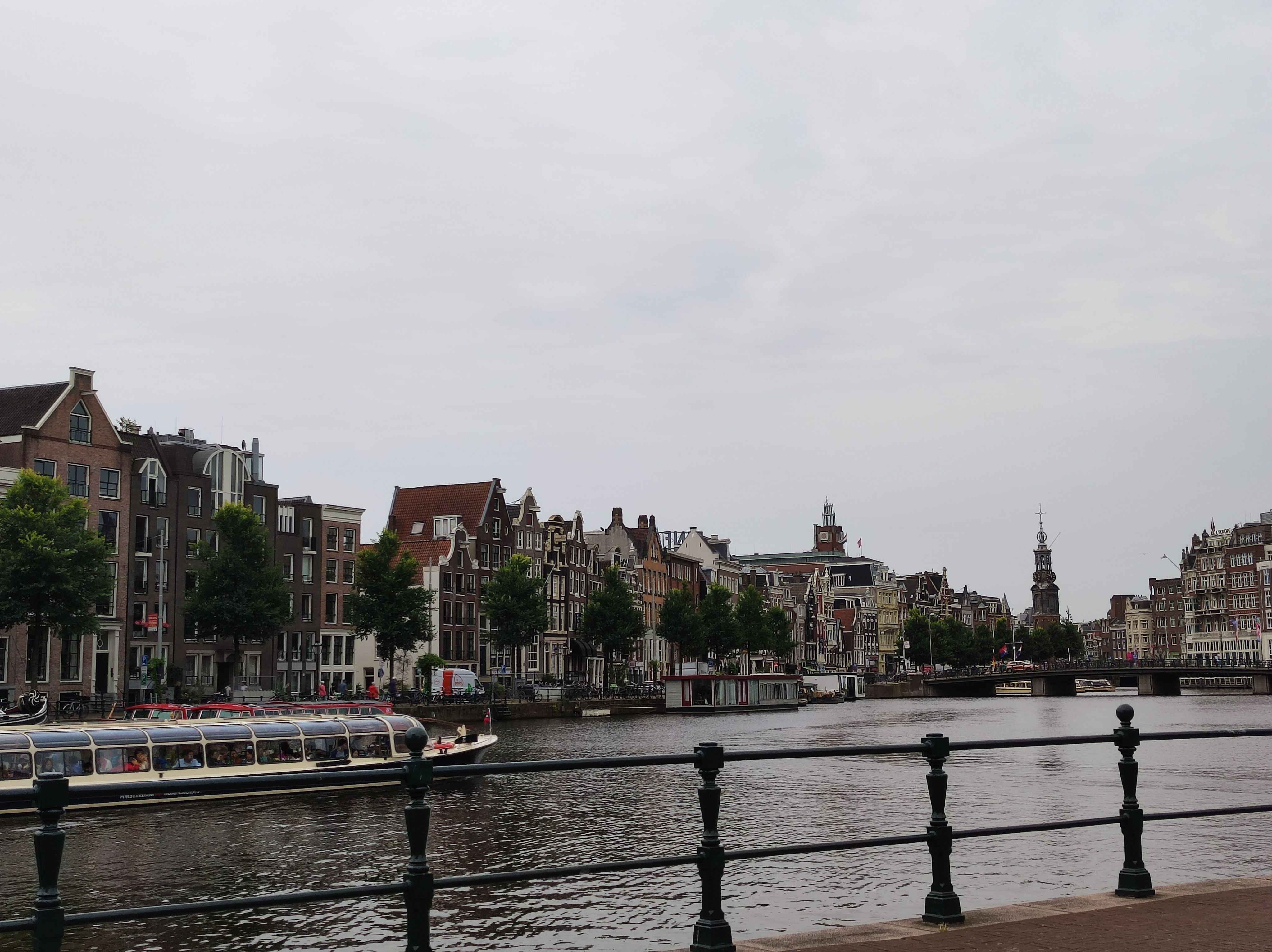Photo of Amsterdam, taken from behind a railing to a wide canal section. Tourist tour boats are passing through on the left. Across the canal, there is a house front of typical, narrow Amsterdam houses. In the background to the right there is also a church tower.