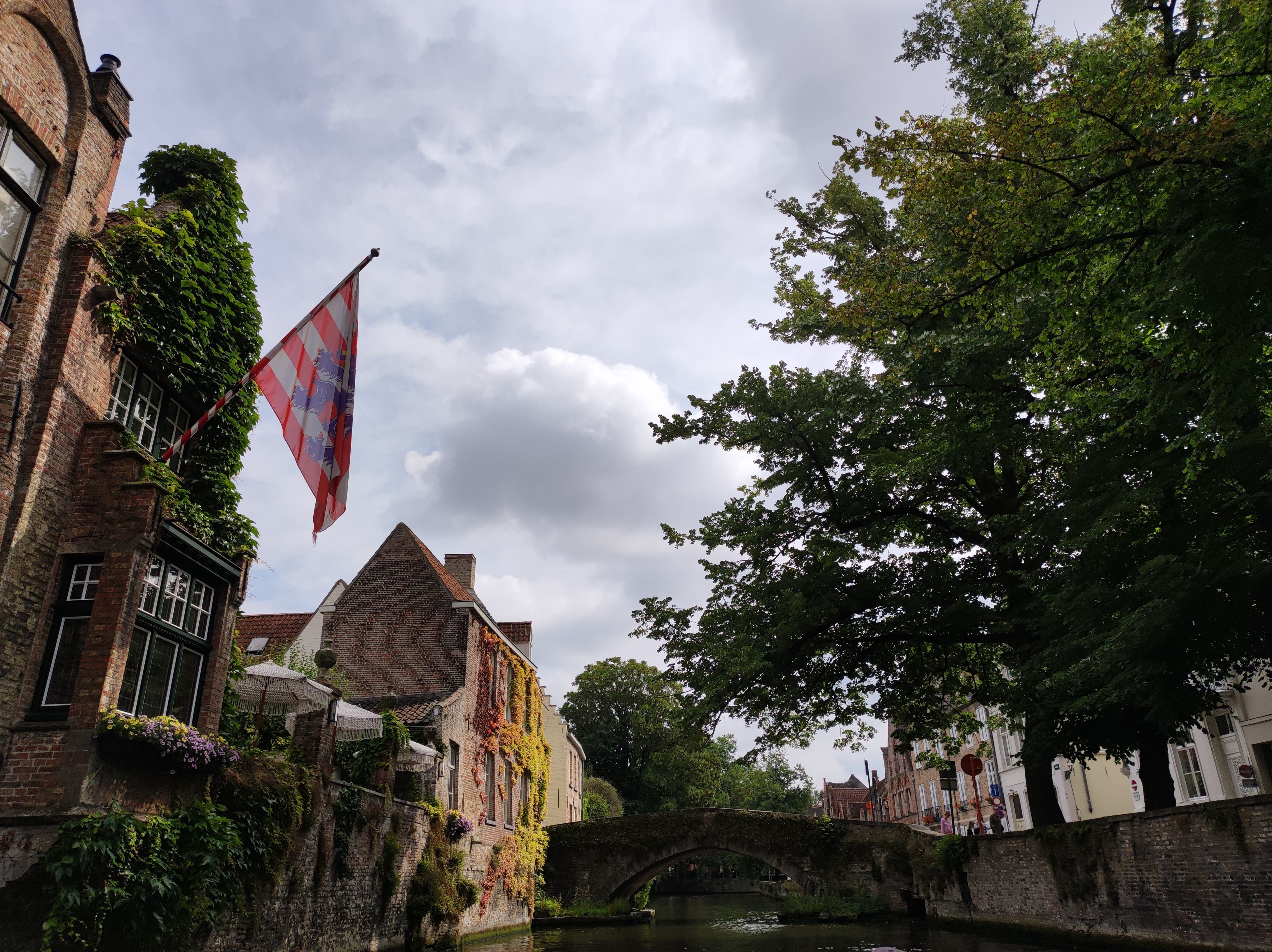 Another photo from the canals of bruges. The view is towards an old stone bridge. To the left, we see brick houses covered in moss and plants, and a terace with sunshades. The house in the foreground displays the Bruges flag.