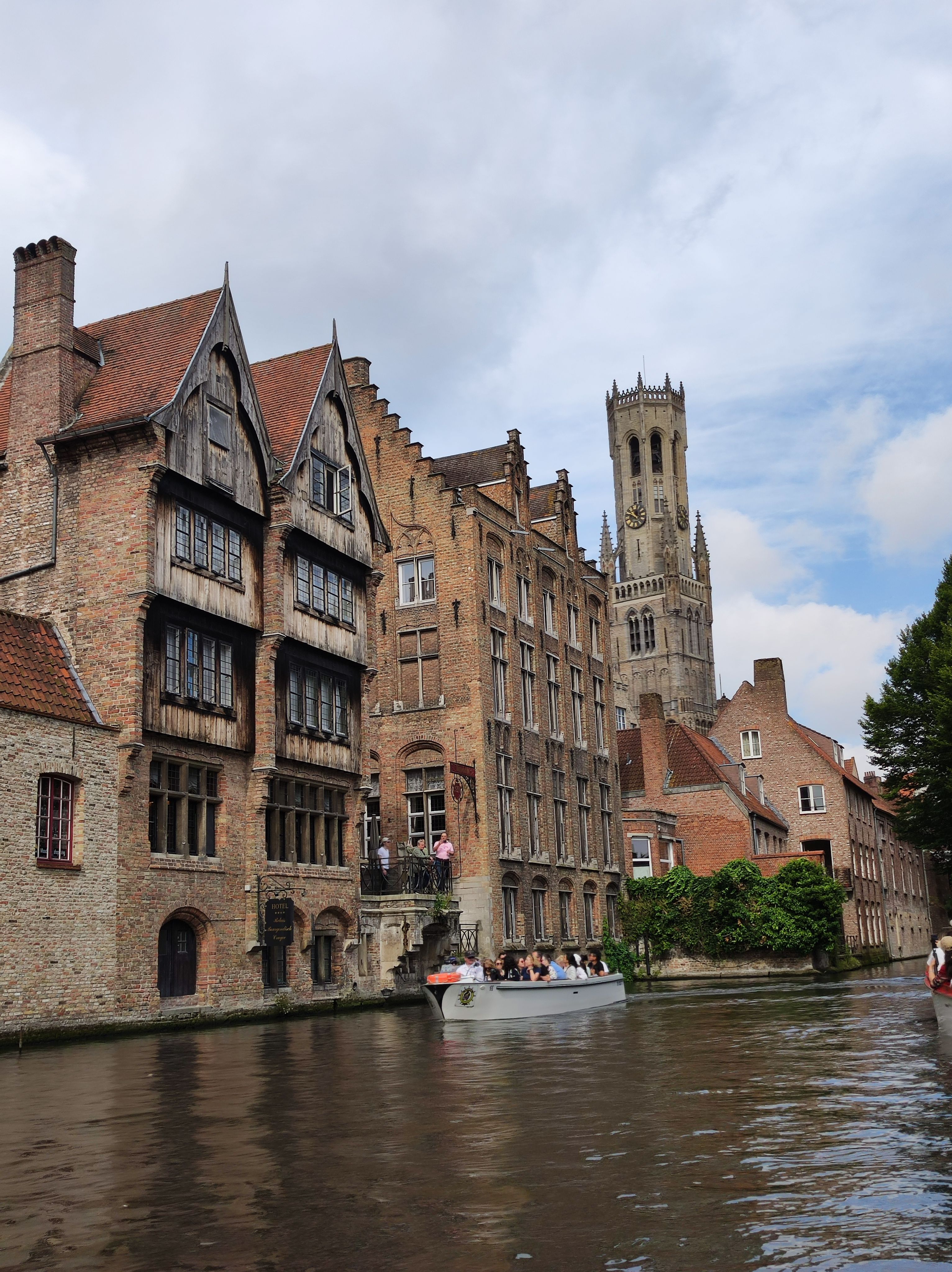 Photo of Bruges from a canal. On the shore to the left, there are a couple old-looking houses, one of them with a wood facade. Further in the background, the Belfort tower is visible over the closer houses. Another boat of tourists is visible on the canal as well.