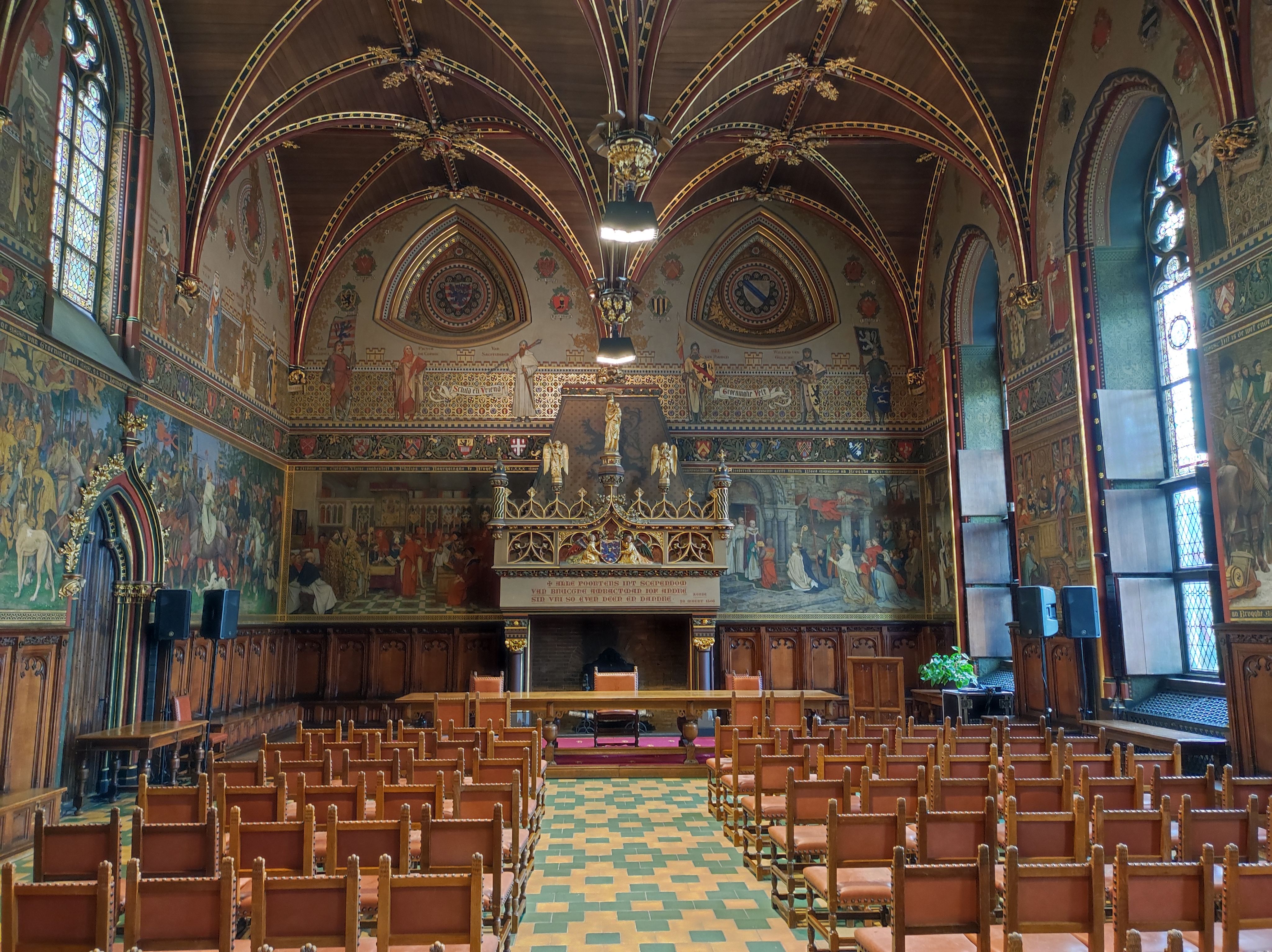 Photo of the interior of the city hall's 'Gothic Hall'. It's a rectangular room with a fireplace at one end, which has a long table and central chair in front of it. Facing that fireplace and table are rows of leather chairs, divided in 2 coloumns. The walls and wood ceiling feature many intrigate ornaments. The walls are also covered in colourful paintings displaying people in many different situations. To the right, sunlight is shining in through 2 tall windows.