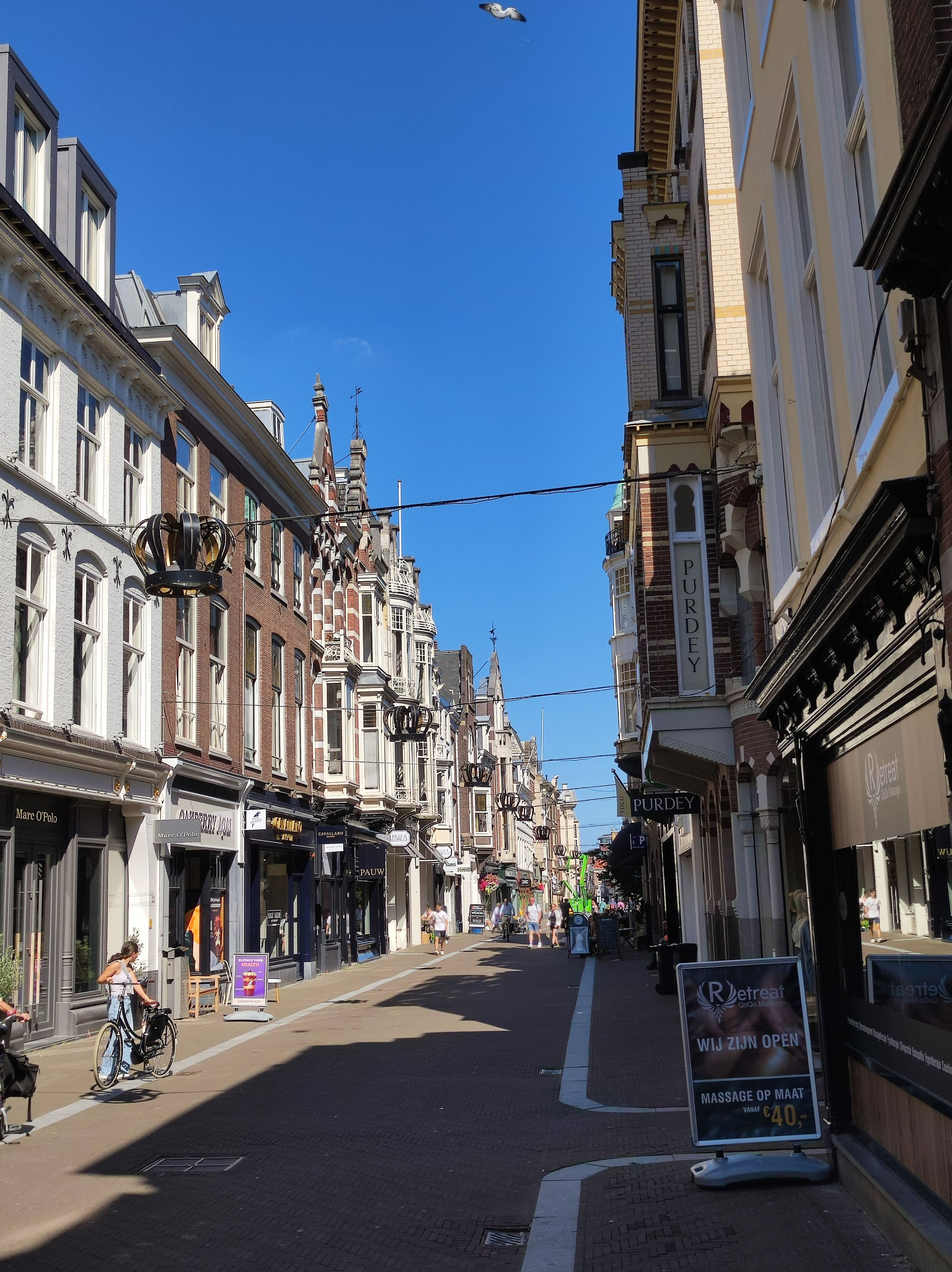 Photo of Noordeinde street in the Hague. A fairly narrow street without cars, many pretty buildings that house different kinds of shops left and right. some pedestrians and cyclists are visible. Between the buildings, metal crowns are suspended on wires.